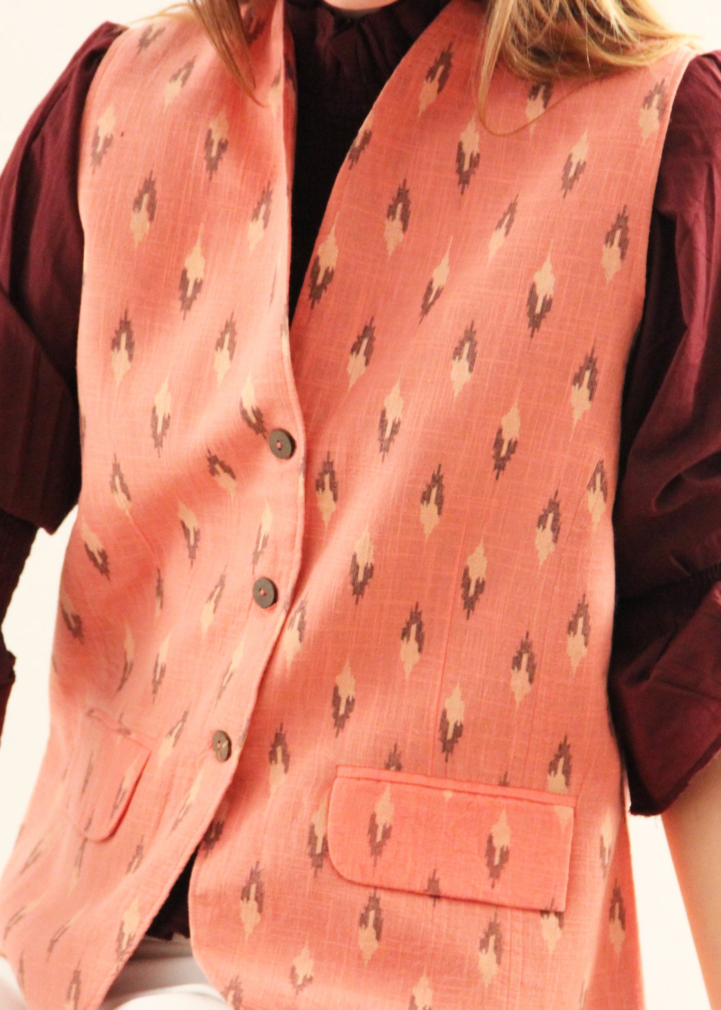 Gilet Madeleine in lino rosa di IGI Ama, smanicato rosa salmone con motivo a rombi, metà rosa chiaro e metà marrone. Realizzato in lino con pattern ikat e interno in velluto a coste. Stile elegante e raffinato, prodotto eticamente in India. Lavabile in lavatrice a 30°C, stirare a bassa temperatura.