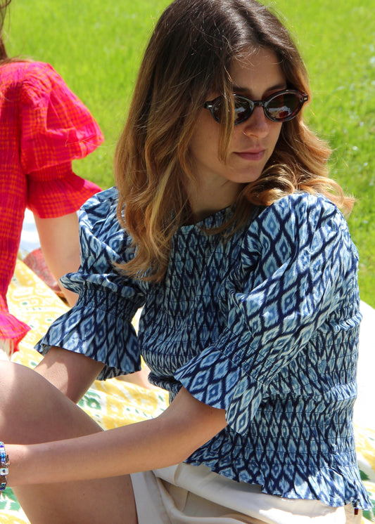 Holi Blouse – Camicetta ikat a maniche corte, realizzata in 100% cotone con maniche a sbuffo e arricciature elastiche su busto e maniche. Vestibilità elasticizzata e confortevole, adatta a tutte le corporature. Taglia unica. Prodotta eticamente in India. Cura delicata: lavare a 30°C, stirare a bassa temperatura.