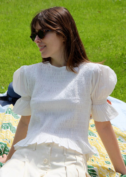 Holi Blouse – Camicetta bianca a maniche corte, realizzata in 100% cotone con maniche a sbuffo e arricciature elastiche su busto e maniche. Vestibilità elasticizzata e confortevole, adatta a tutte le corporature. Taglia unica. Prodotta eticamente in India. Cura delicata: lavare a 30°C, stirare a bassa temperatura.